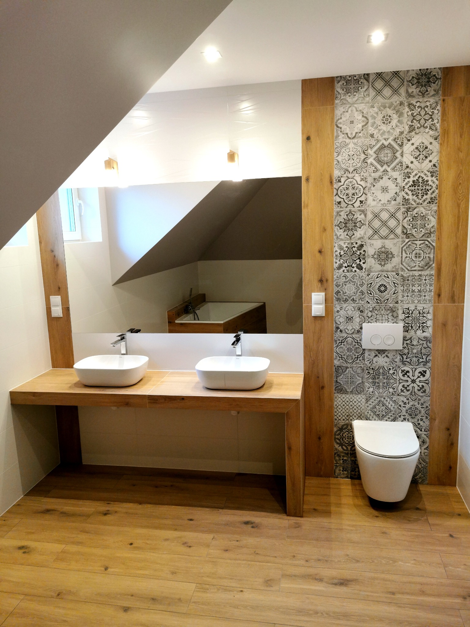 Nowoczesna łazienka na poddaszu z drewnopodobną podłogą i blatem pod umywalki, dekoracyjnymi płytkami na ścianie oraz wiszącą toaletą.