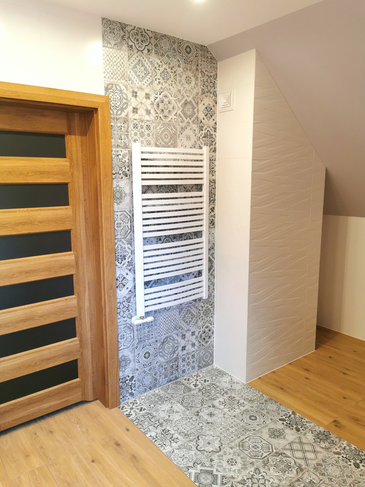 Wykończona łazienka w stylu patchwork z drewnianą podłogą i drzwiami, grzejnik drabinkowy na ścianie z wzorzystych płytek.