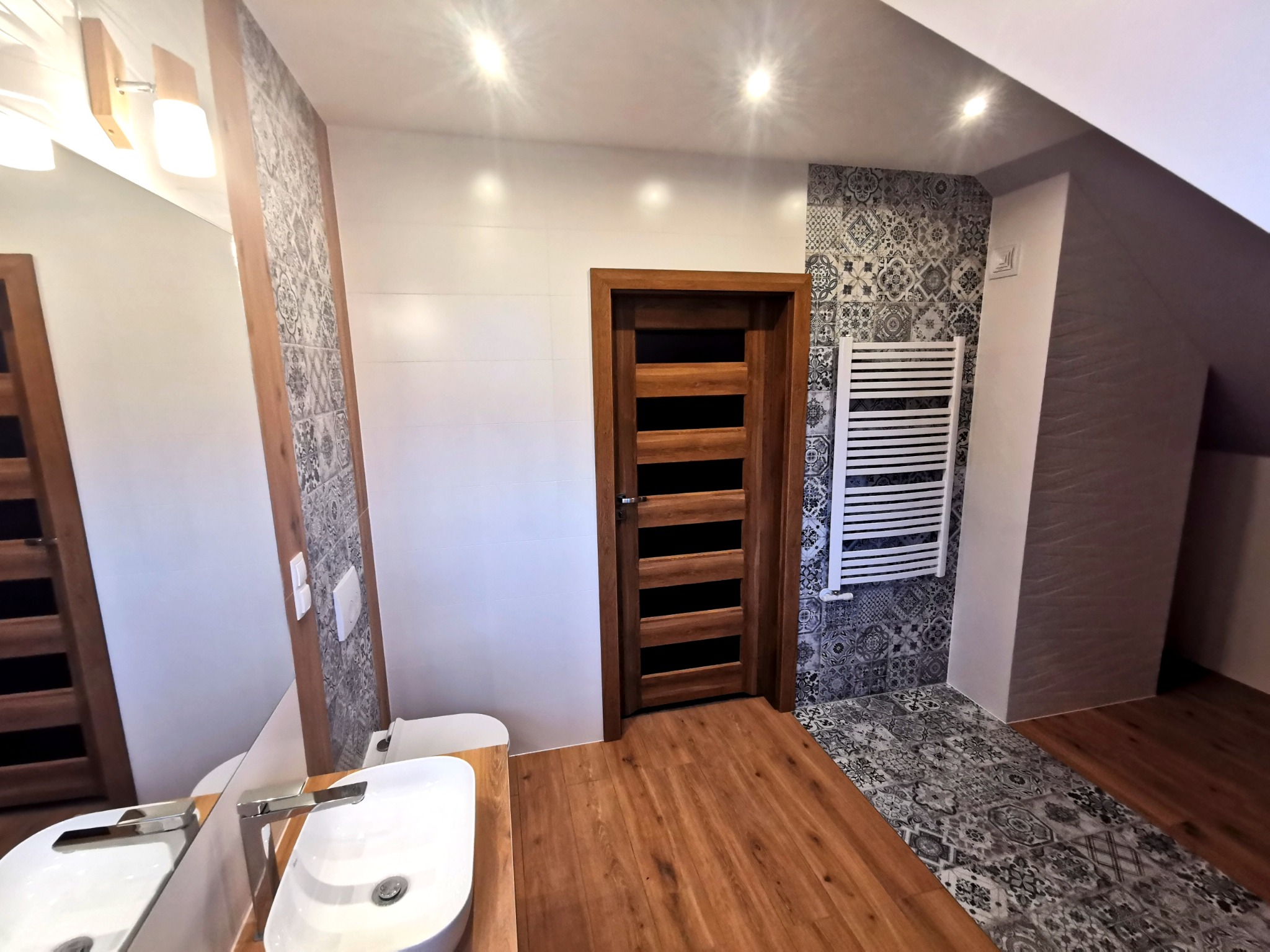 Nowoczesna łazienka na poddaszu z drewnianą podłogą, umywalką nablatową, lustrem z oświetleniem, grzejnikiem drabinkowym i wzorzystymi płytkami w strefie prysznica.