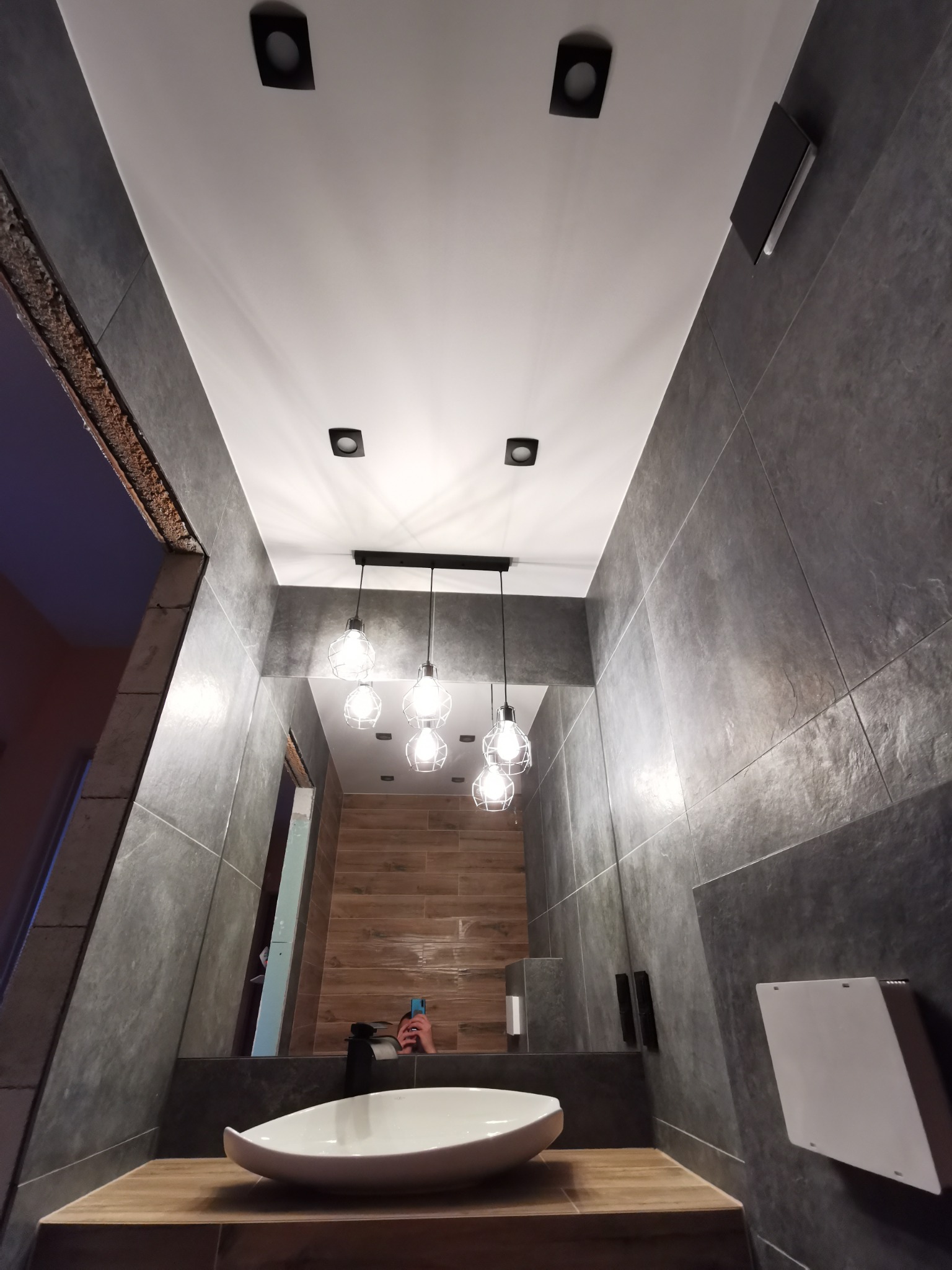 Nowoczesna łazienka z szarymi płytkami, drewnianym blatem pod umywalką i wiszącymi lampami z żarówkami Edison, widok z dołu.