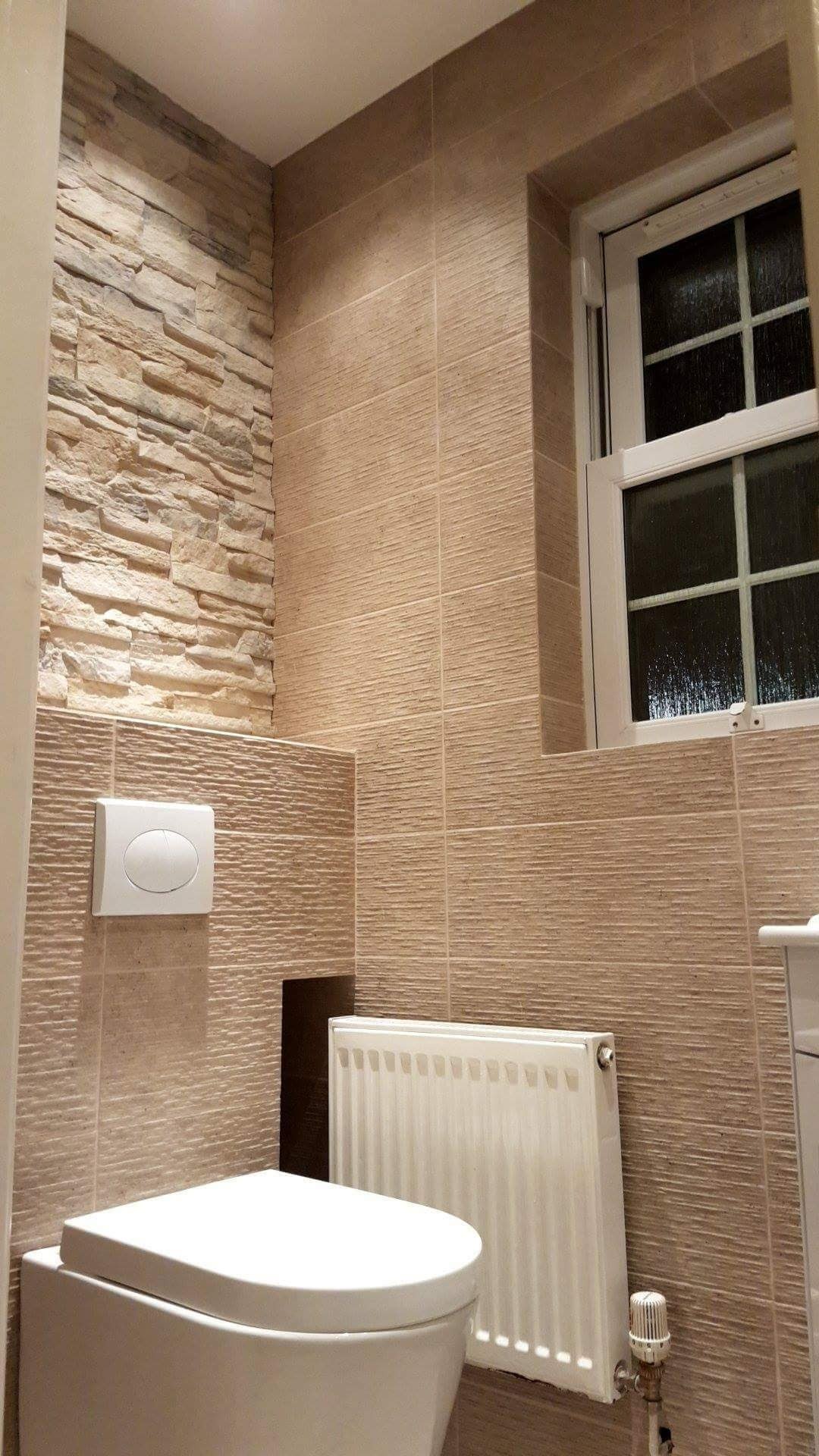 Wykończona łazienka z beżowymi płytkami imitującymi kamień na ścianach, biała toaleta z podwieszanym zbiornikiem, biały grzejnik i okno z białymi ramami.