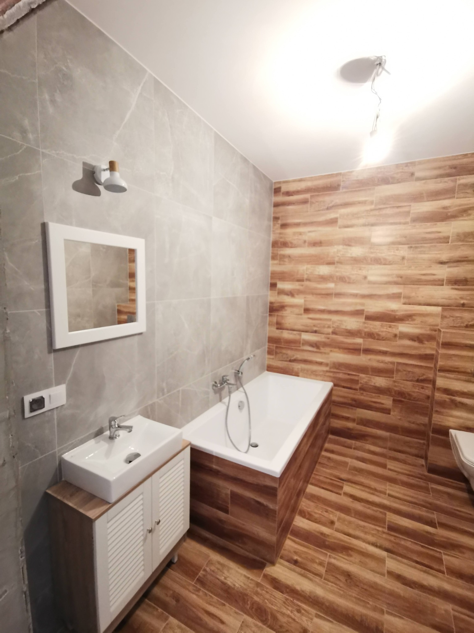 Wykończona łazienka z szarymi płytkami imitującymi marmur na ścianach i drewnopodobnymi płytkami na podłodze i jednej ze ścian, biała wanna obudowana drewnopodobnymi panelami, biała szafka...