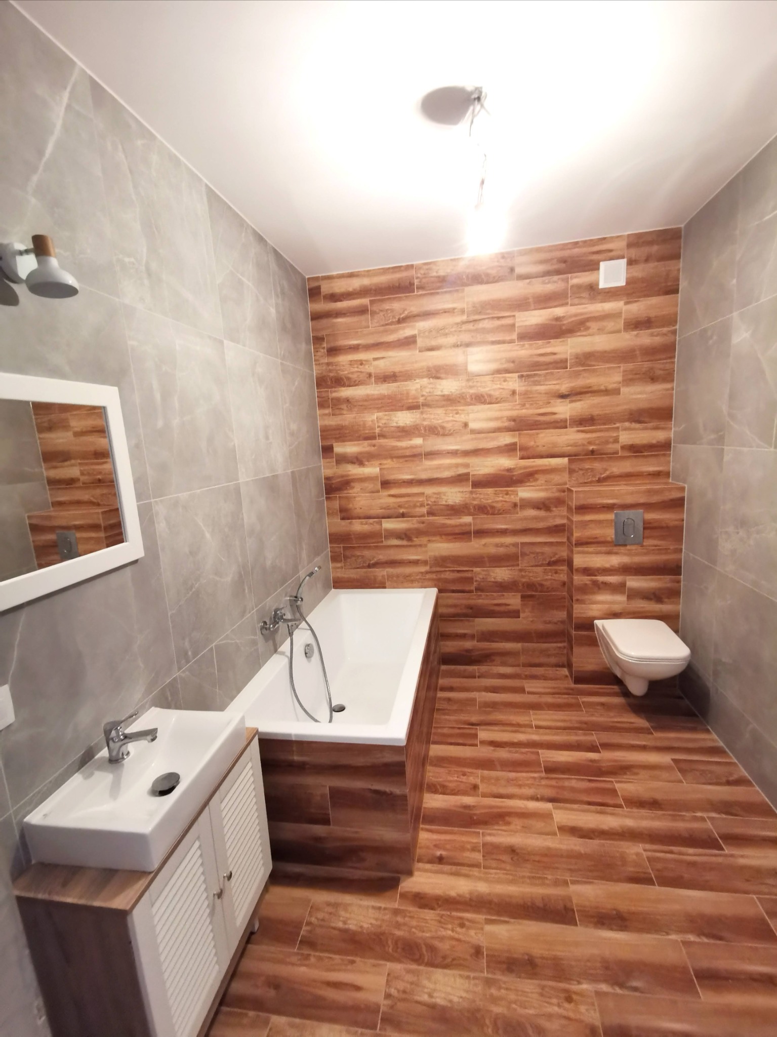Wykończona łazienka z drewnopodobnymi płytkami na podłodze i ścianie za toaletą oraz wanną, szare płytki na pozostałych ścianach, biała umywalka z szafką i lustrem.