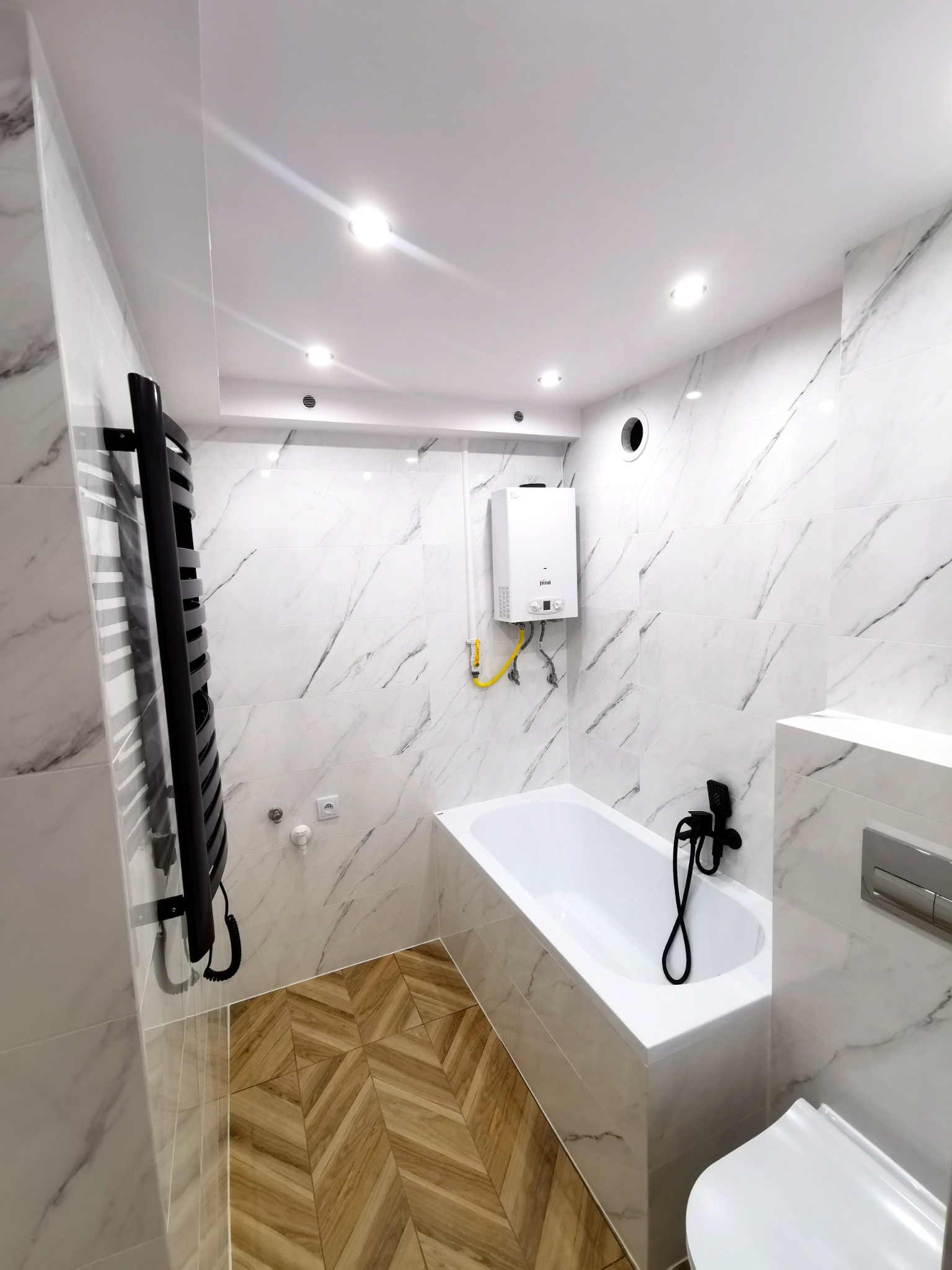 Nowoczesna łazienka z białymi płytkami imitującymi marmur, drewnianą podłogą ułożoną w jodełkę, białą wanną, czarną baterią wannową i grzejnikiem drabinkowym w czarnym kolorze, a także białym...