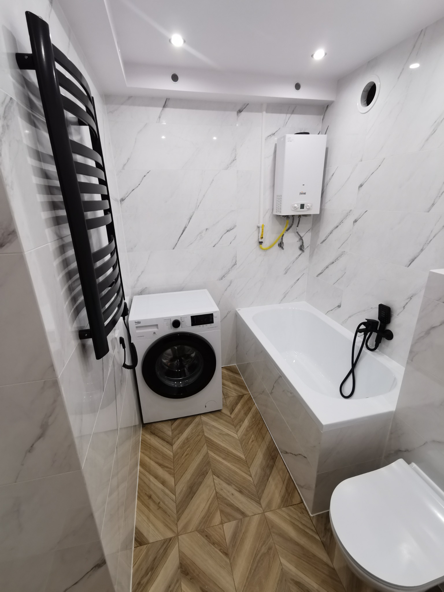 Nowoczesna, jasna łazienka z marmurowymi płytkami, drewnopodobną podłogą w jodełkę, białą wanną z czarną armaturą, pralką Beko i czarnym grzejnikiem drabinkowym.