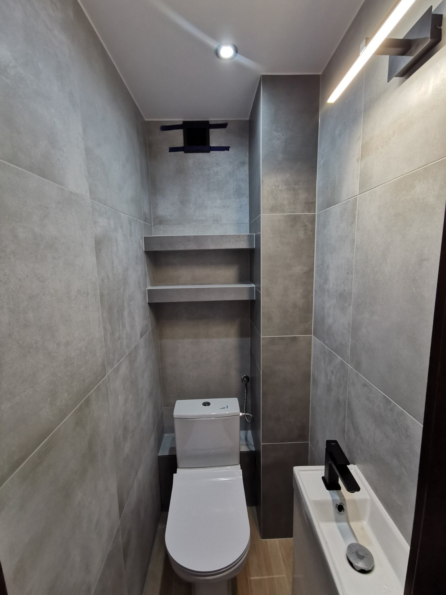 Nowoczesna łazienka w odcieniach szarości z wiszącą toaletą, umywalką z czarną armaturą, półkami ściennymi i oświetleniem LED.