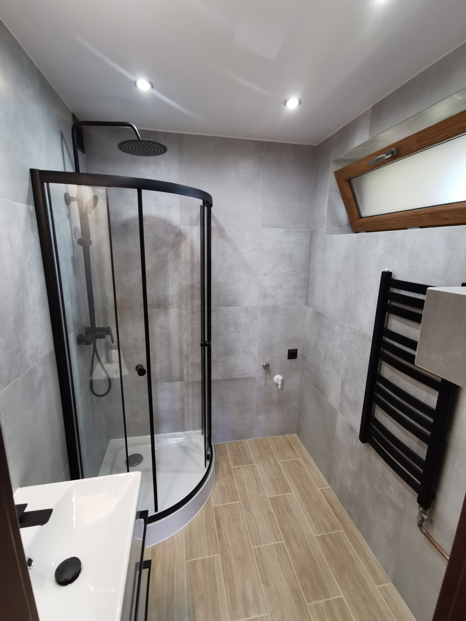 Nowoczesna łazienka z kabiną prysznicową z czarnymi ramami, grzejnikiem drabinkowym w kolorze czarnym, umywalką z czarną armaturą, szarymi płytkami ściennymi i podłogą imitującą drewno.