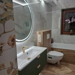 PawBud - Nowoczesna łazienka z umywalką w szafce w kolorze butelkowej zieleni, okrągłym lustrem z podświetleniem LED, płytkami imitującymi marmur i drewnopodobną podłogą oraz wiszącą toaletą.