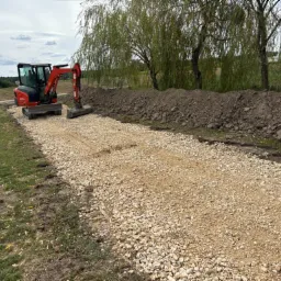 Czerwony mini-koparka Kubota na wysypanym tłuczniem terenie, obok pryzma ziemi, w tle drzewa i niebo z chmurami.