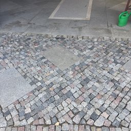 PawBud - Starannie ułożona kostka brukowa w odcieniach szarości i różu, tworząca diagonalny wzór, połączona z szarymi płytami granitowymi z jasnym prostokątnym wkładem. W tle zielona konewka.