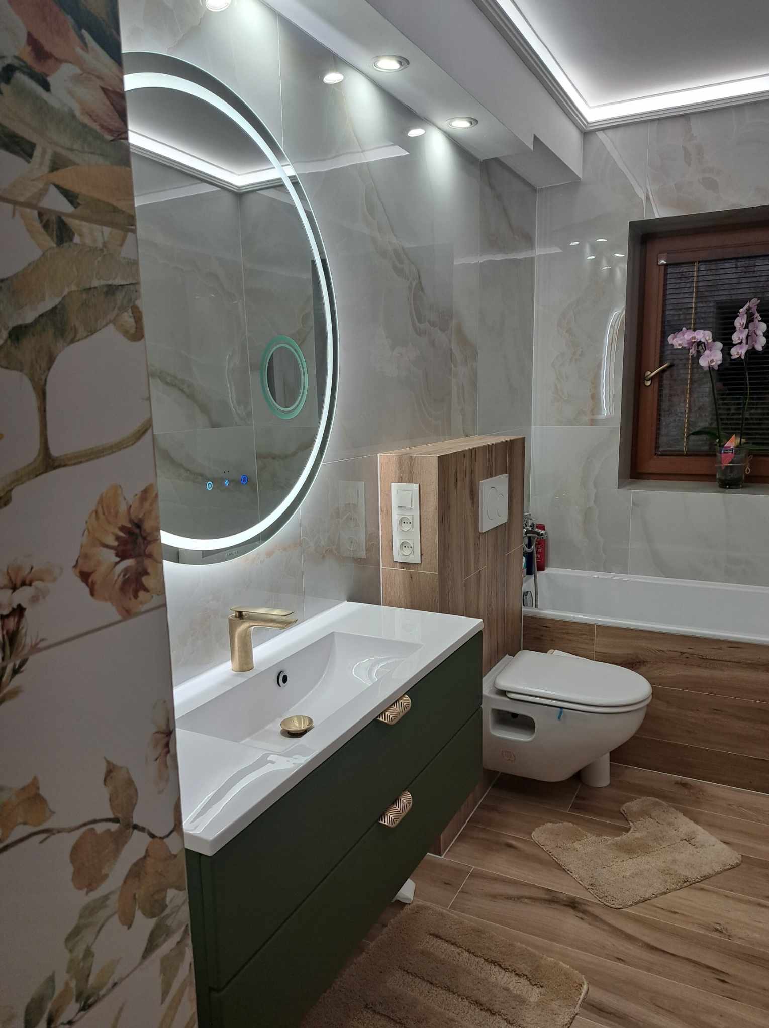 Nowoczesna łazienka z umywalką w szafce w kolorze butelkowej zieleni, okrągłym lustrem z podświetleniem LED, płytkami imitującymi marmur i drewnopodobną podłogą oraz wiszącą toaletą.