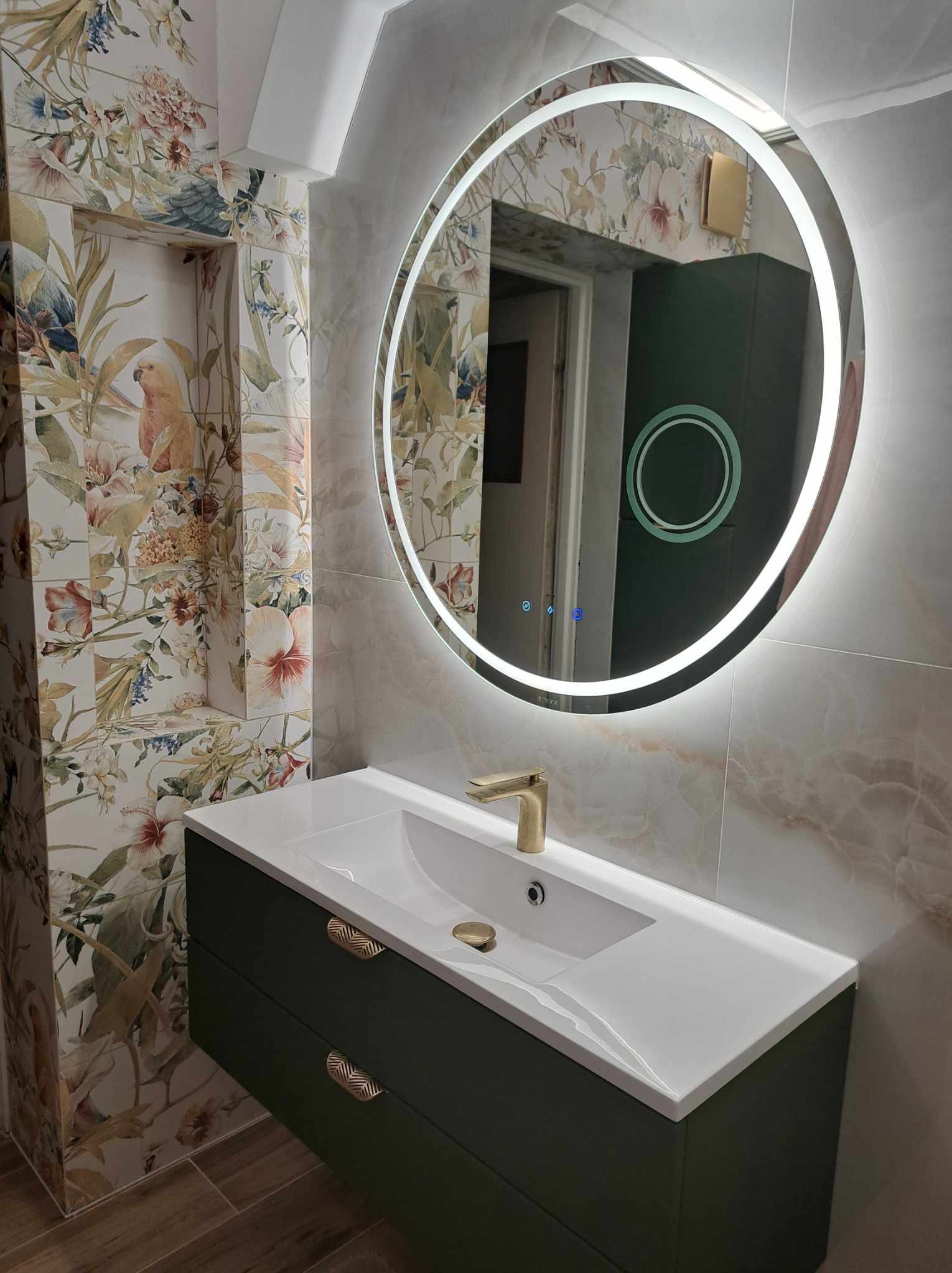 Nowoczesna łazienka w Staszowie z umywalką nablatową, szafką w kolorze butelkowej zieleni i okrągłym lustrem z podświetleniem LED, na ścianach płytki w kwiatowe wzory i imitujące marmur.