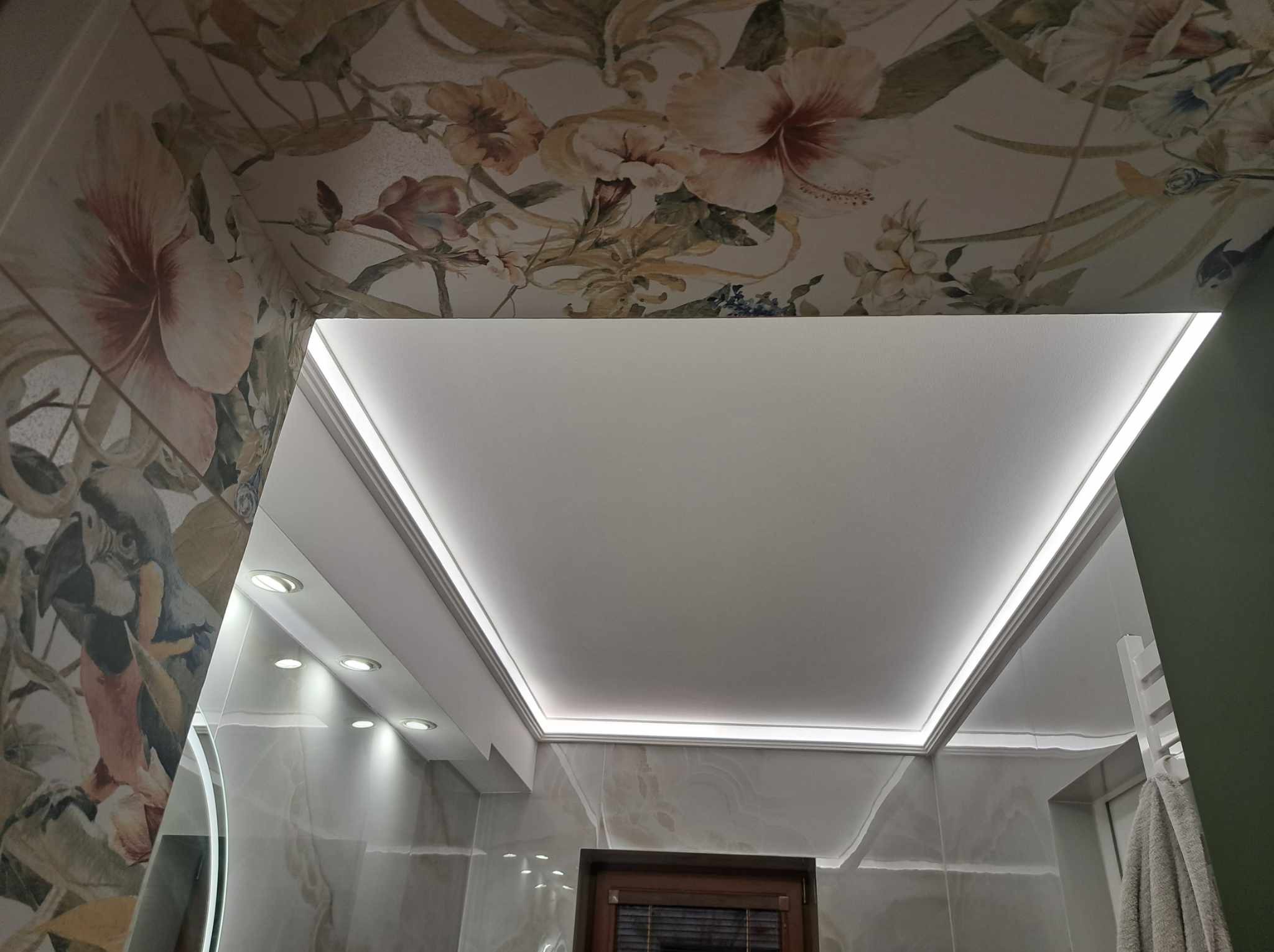 Podwieszany sufit z oświetleniem LED w łazience z tapetą w kwiaty i motywy tropikalne na ścianach i suficie.