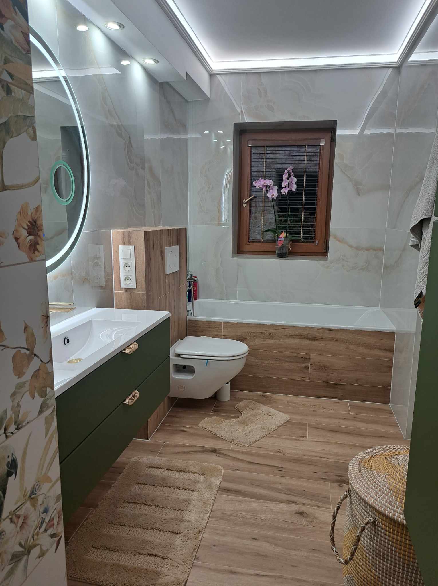 Nowoczesna łazienka z drewnopodobnymi płytkami, zieloną szafką pod umywalką z dekoracyjnymi uchwytami, okrągłym lustrem z podświetleniem LED i drewnianą ramą okienną z orchideą.