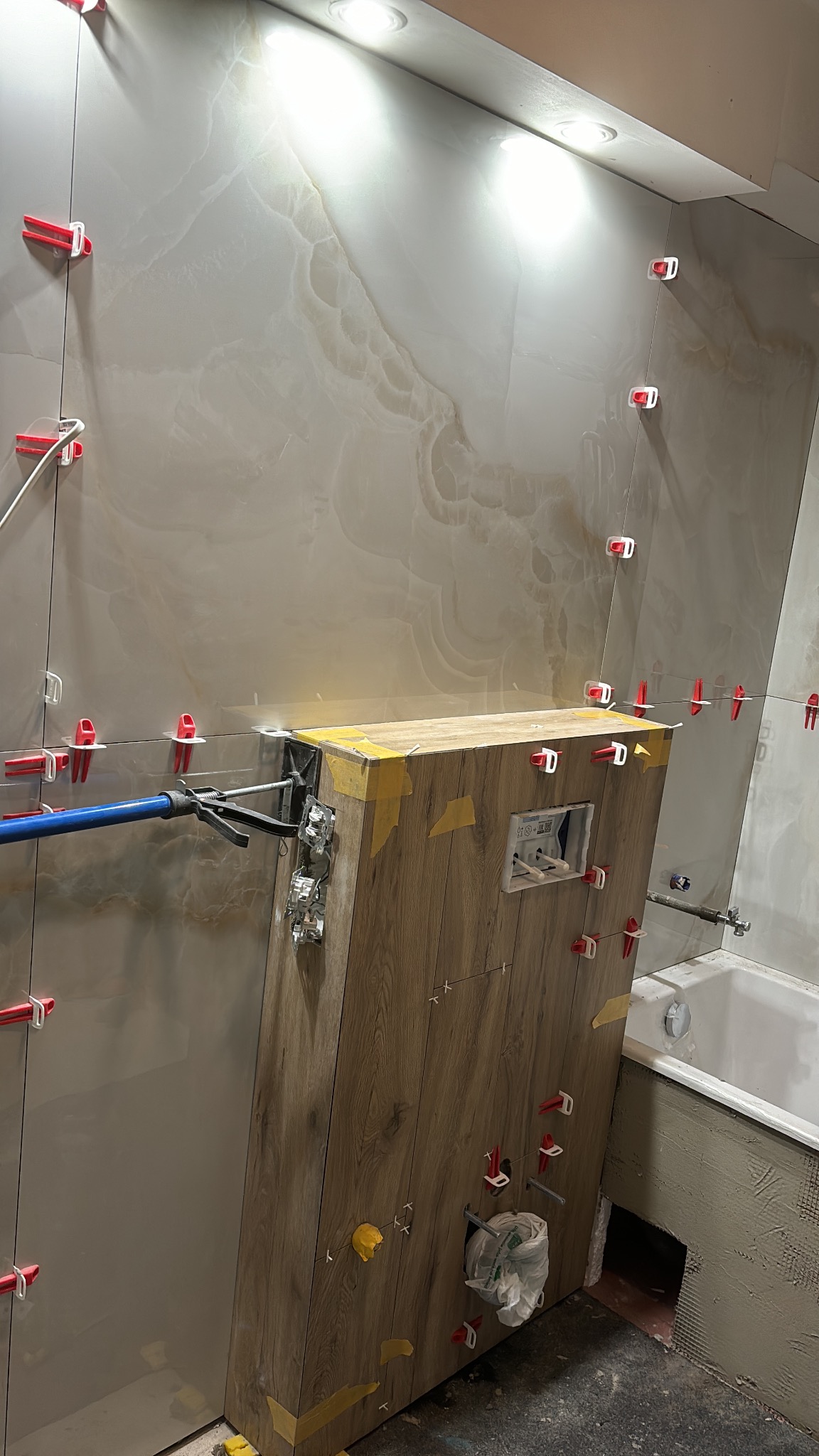 Montaż płytek imitujących marmur w łazience, widoczne krzyżyki dystansowe i fragment zabudowy stelaża WC obłożonego płytkami drewnopodobnymi, z widocznymi otworami na instalacje.