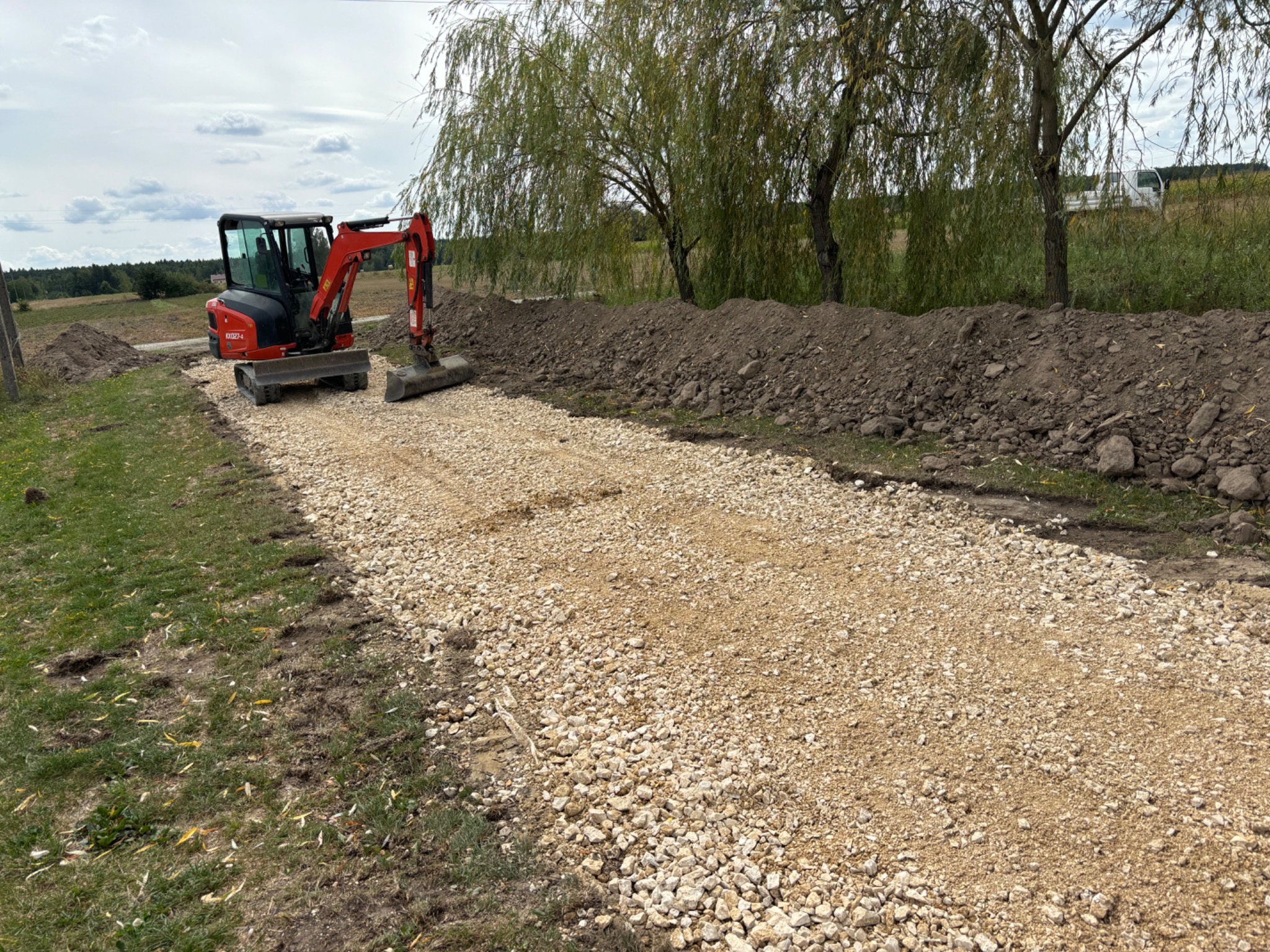 Czerwony mini-koparka Kubota na wysypanym tłuczniem terenie, obok pryzma ziemi, w tle drzewa i niebo z chmurami.