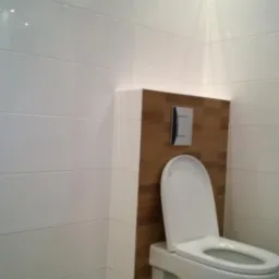 Nowoczesna toaleta podwieszana z drewnopodobną obudową stelaża i białymi, poziomymi płytkami na ścianach oraz drewnianą podłogą, oświetlona punktowo.