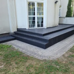 Czarny, wielopoziomowy taras z kompozytu na zewnątrz budynku, z widocznymi schodami i obrzeżem z kostki brukowej, przylegający do białej ściany z oknami.