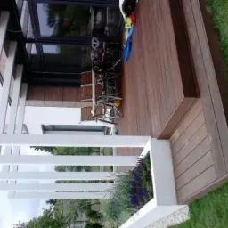 Nowoczesny taras z desek w odcieniach brązu, z białymi elementami konstrukcyjnymi, otoczony zielonym trawnikiem i niską roślinnością.