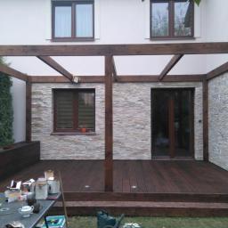 Korsuper - Drewniana pergola z zadaszeniem nad tarasem z desek, widoczna lampa na ścianie, w tle dom z oknami i kamienną elewacją, na pierwszym planie stół z narzędziami i akcesoriami.