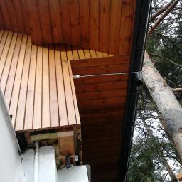 Korsuper - Fragment elewacji domu z drewnianą okładziną, widoczne ocieplenie i rynna, na tle drzewa iglastego.
