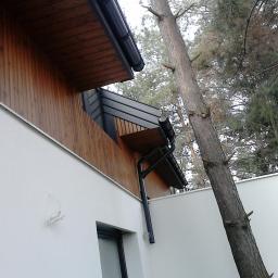 Korsuper - Fragment elewacji domu z drewnianą okładziną i białym tynkiem, widoczne rynny, drzewo iglaste w tle.