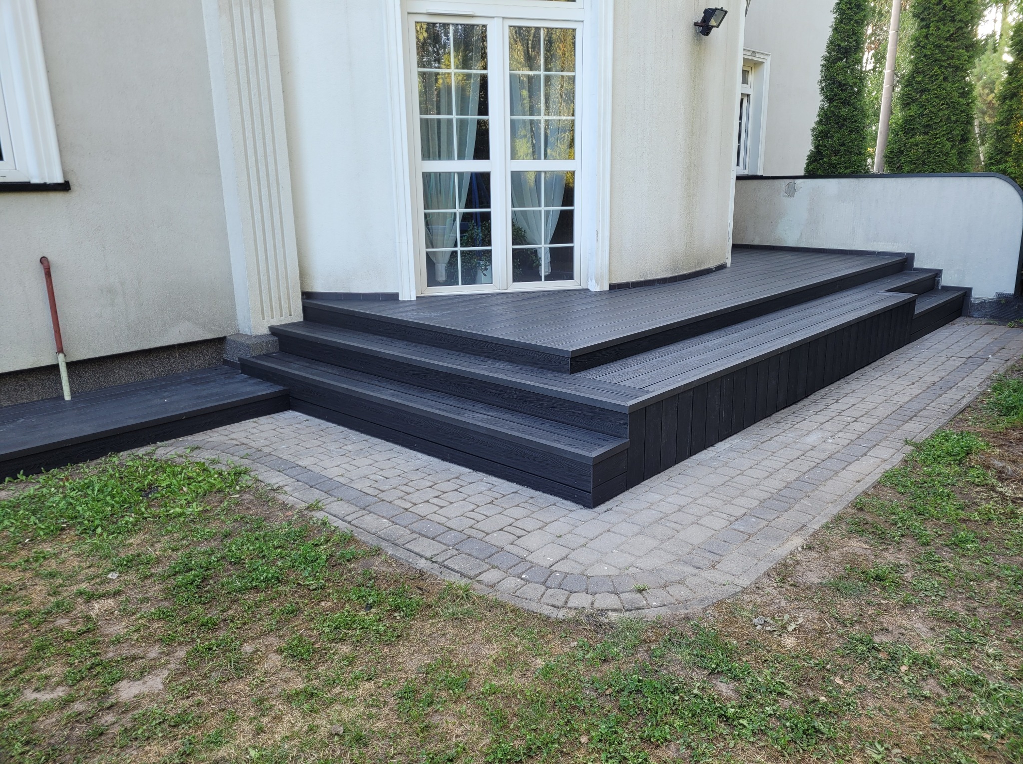 Czarny, wielopoziomowy taras z kompozytu na zewnątrz budynku, z widocznymi schodami i obrzeżem z kostki brukowej, przylegający do białej ściany z oknami.