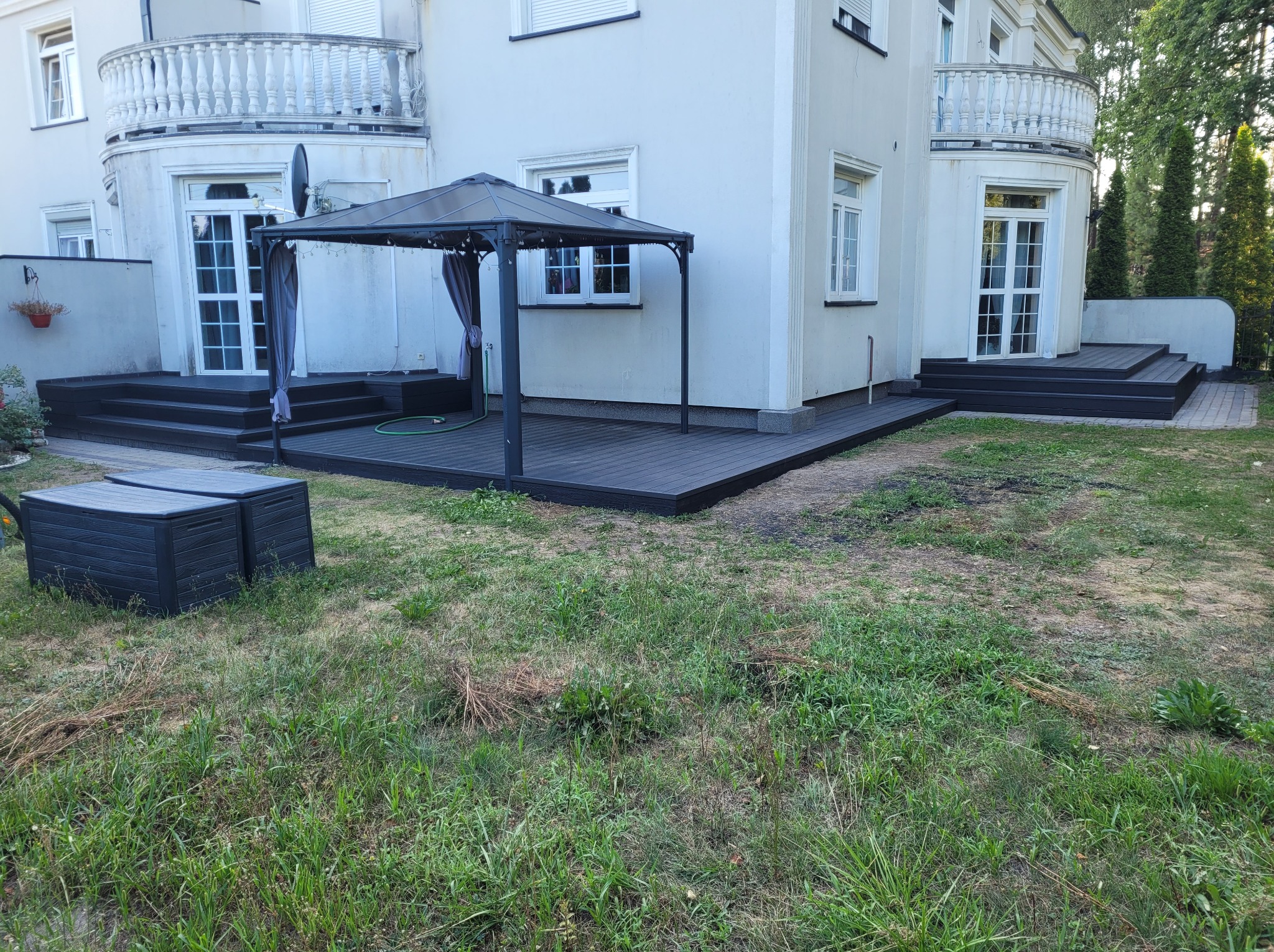 Ciemny taras z kompozytu zadaszony altaną przy białym domu z balkonami, dwa pojemniki na trawie z przodu.