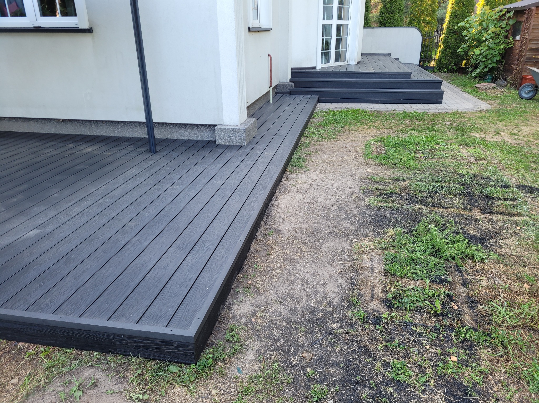 Nowoczesny, ciemnoszary taras z desek kompozytowych przylegający do białego domu z widocznymi schodami i fragmentem trawnika z chwastami.