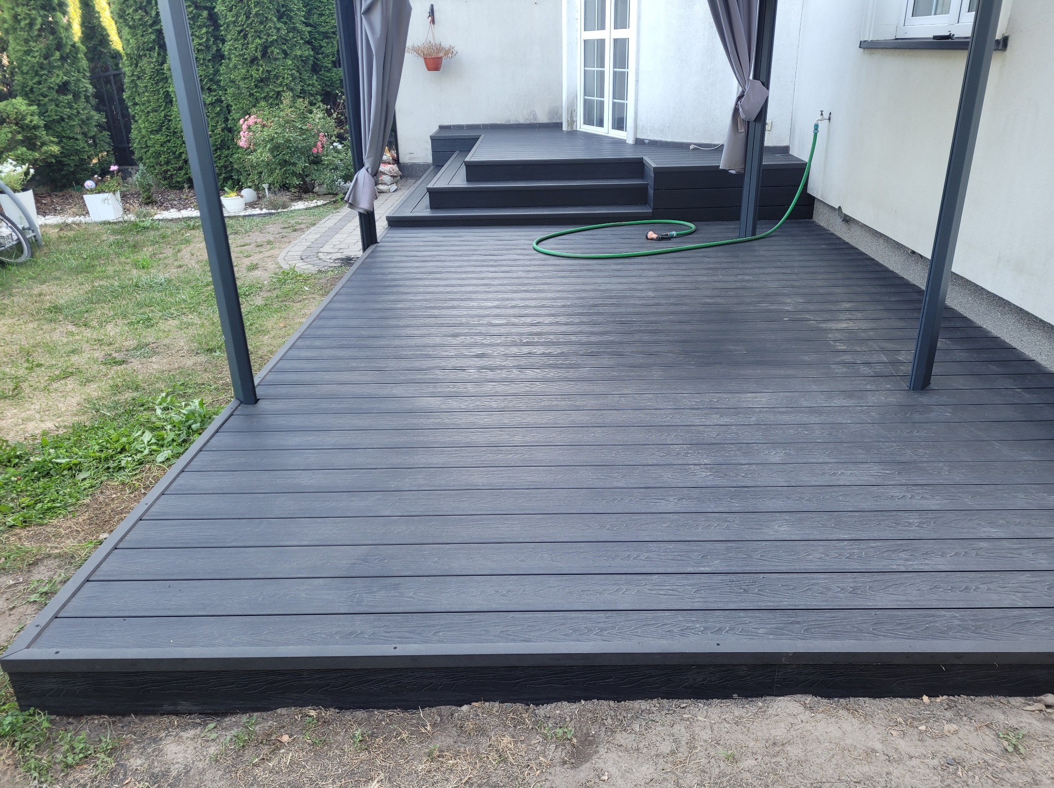 Nowoczesny taras z szarych desek kompozytowych, zadaszony pergolą z szarymi zasłonami, prowadzący do drzwi wejściowych, z zielonym wężem ogrodowym na powierzchni.