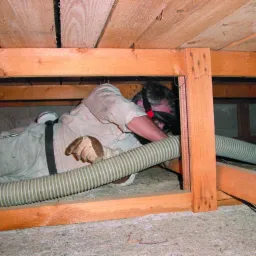 Pracownik w kombinezonie ochronnym i masce, leżąc w przestrzeni poddasza, aplikuje izolację za pomocą węża, widoczne drewniane elementy konstrukcyjne dachu i ściany.