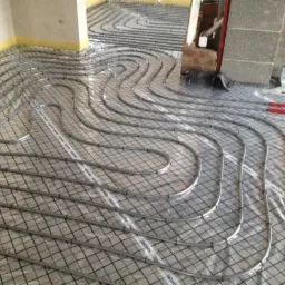 Ułożone rury ogrzewania podłogowego na folii z siatką, widok z góry. Fragment ściany z żółtą taśmą i elementy konstrukcyjne w tle.