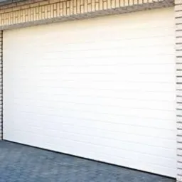 Biała segmentowa brama garażowa z poziomymi panelami, widoczna od frontu, z fragmentem ceglanej ściany po bokach i brukowaną posadzką u dołu.