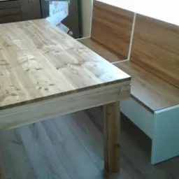 Drewniany stół z ławką narożną w jasnej kolorystyce, z widoczną strukturą drewna, ustawiony na panelach podłogowych.