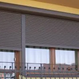 Brązowa roleta zewnętrzna opuszczona na okno, widoczne lamele i kaseta rolety, zarys budynku w tle.