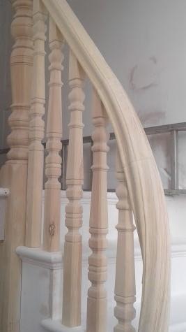 Surowe drewniane poręcze schodowe z ozdobnymi toczonymi słupkami, widoczne ślady obróbki, tło z widoczną drabiną.