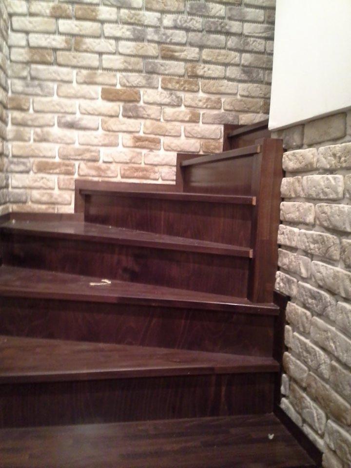 Ciemnobrązowe schody z drewna, z widoczną strukturą słojów, przylegają do ściany wyłożonej dekoracyjną cegłą w odcieniach beżu i szarości.