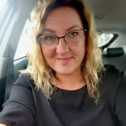 Portret kobiety w okularach z blond włosami, siedzącej w samochodzie. Uśmiechnięta twarz, czarna bluzka, wnętrze pojazdu w tle.