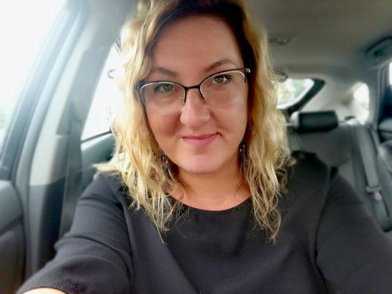 Portret kobiety w okularach z blond włosami, siedzącej w samochodzie. Uśmiechnięta twarz, czarna bluzka, wnętrze pojazdu w tle.