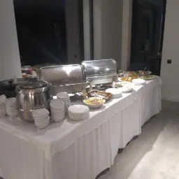 Stół cateringowy przykryty białym obrusem z metalowymi bemarami, naczyniami i potrawami, ustawiony w jasnym wnętrzu z dużym oknem.