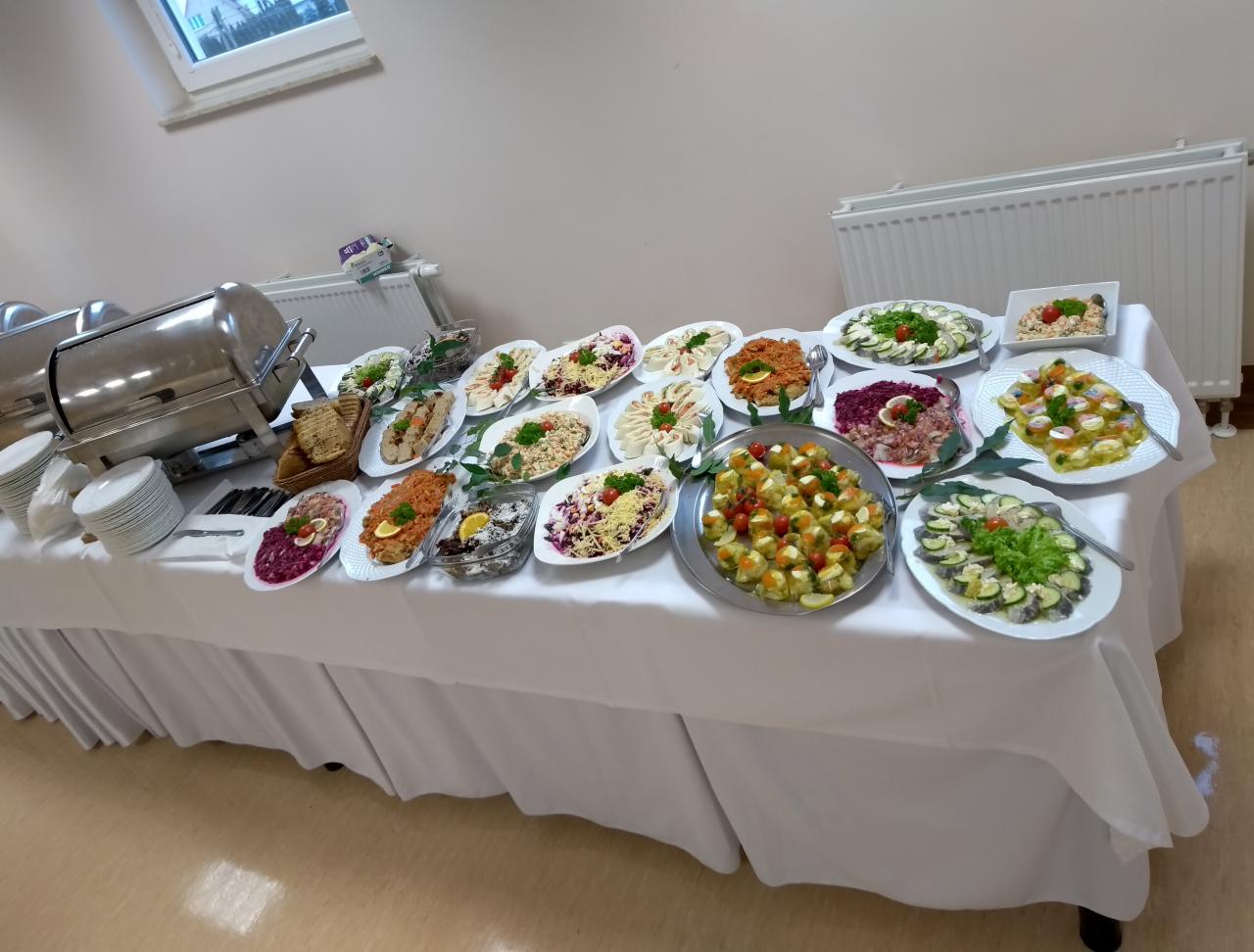 Bufet z różnorodnymi przystawkami: sałatki, koreczki, pieczywo, dania rybne, serwowane na białym obrusie, z widocznymi podgrzewaczami i stosem talerzy.