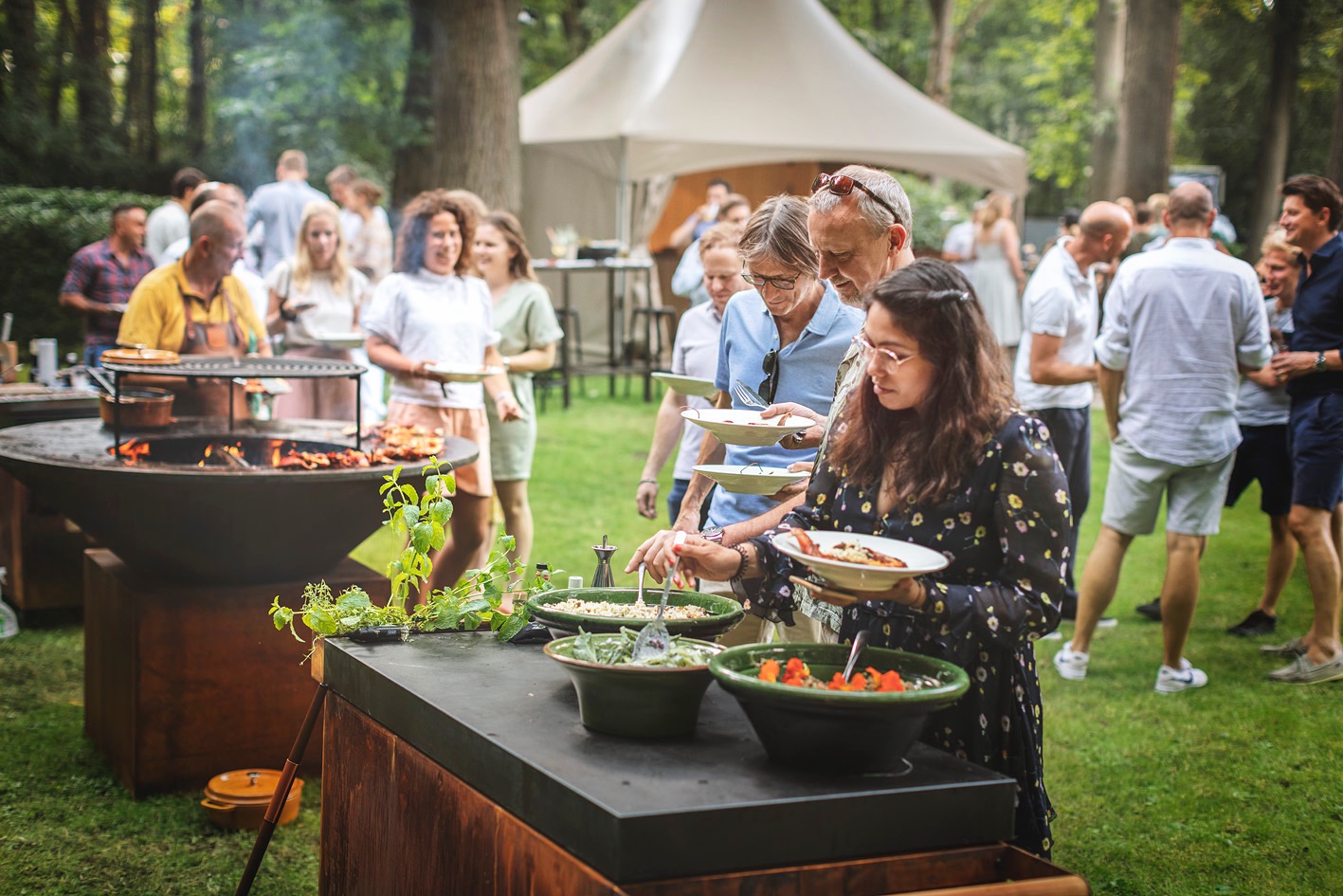 Uczestnicy imprezy plenerowej ustawiają się w kolejce do bufetu z grillem i sałatkami, widoczne płonące drewno w palenisku grilla i zielone otoczenie.