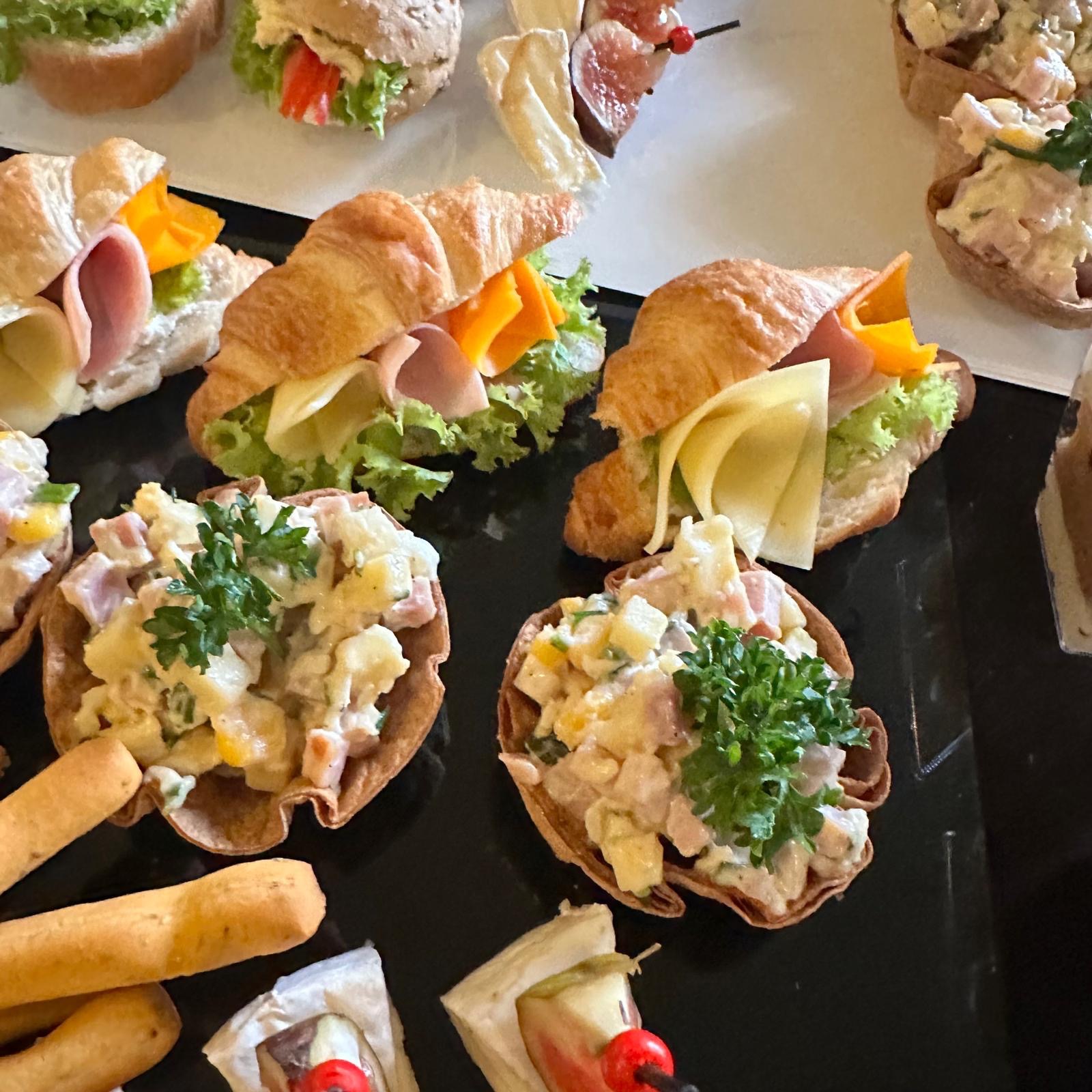 Elegancko ułożone przekąski na czarnym talerzu: mini croissanty z szynką i serem, sałatki w jadalnych koszyczkach, koreczki z figami i roladki z szynką.