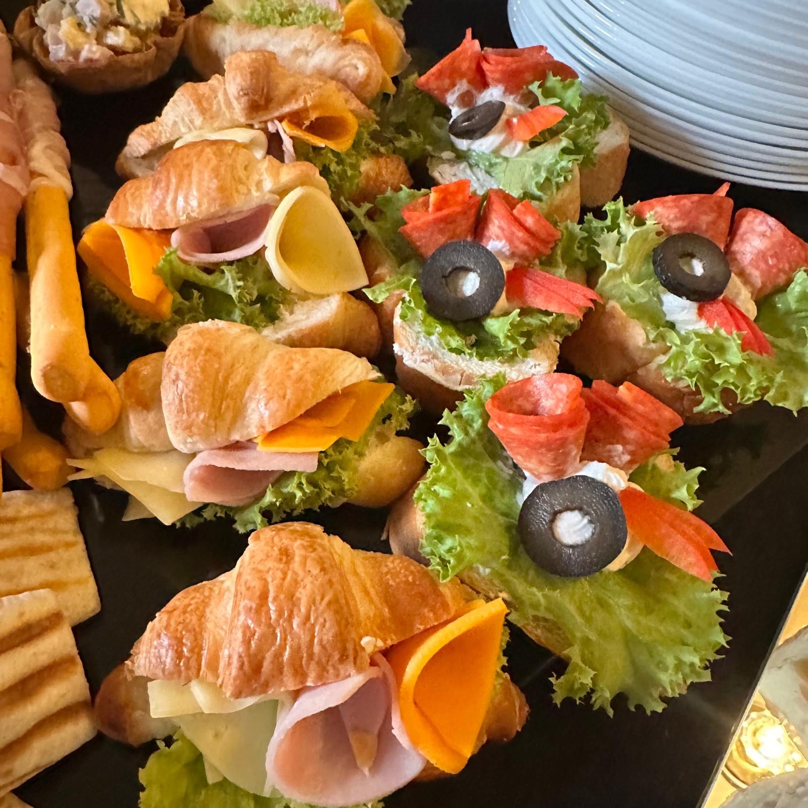 Dekoracyjne przekąski cateringowe: mini croissanty z szynką i serem oraz kanapki z salami, oliwkami i sałatą na czarnym talerzu obok stosu białych talerzy.