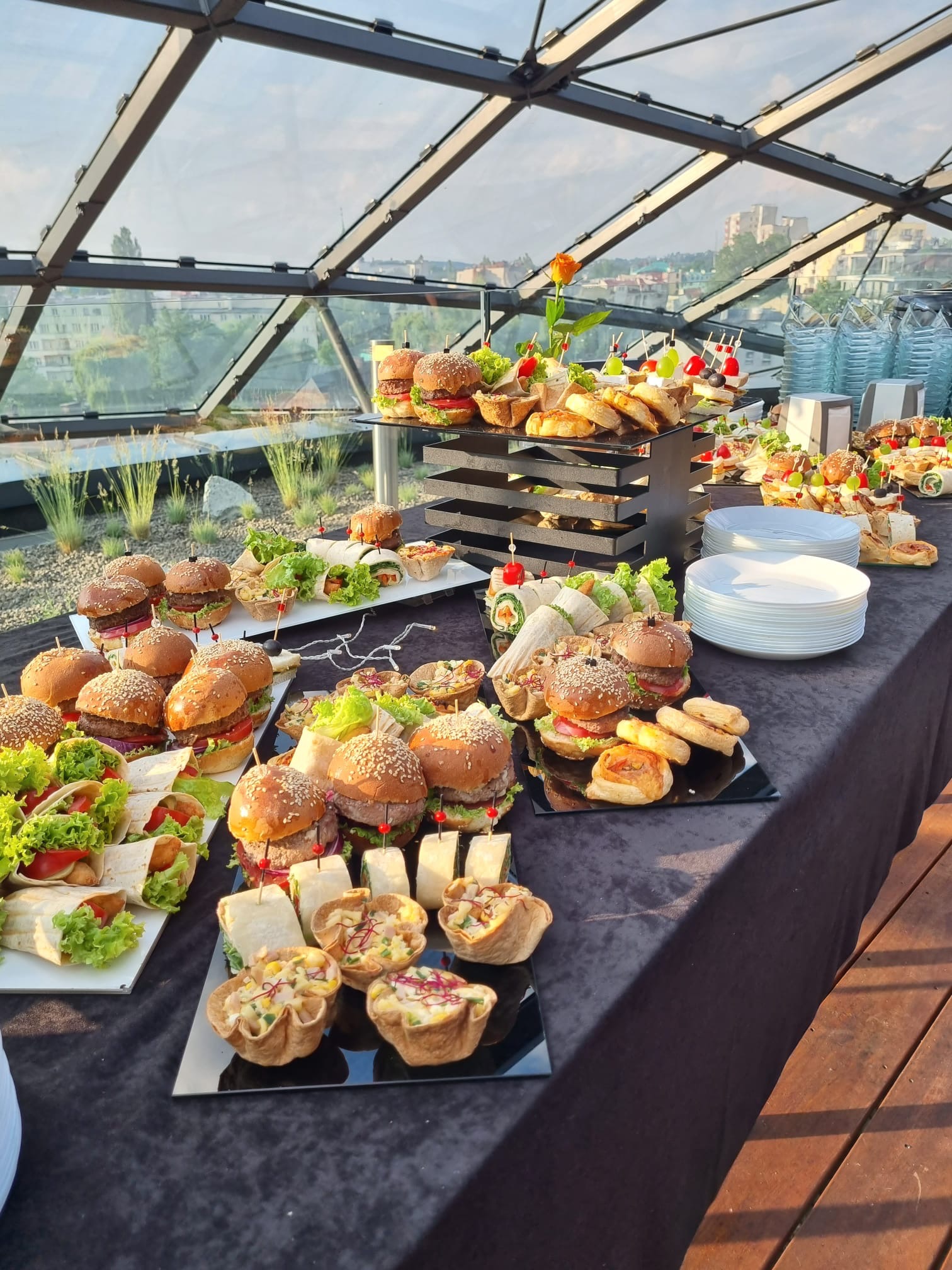 Stół cateringowy z burgerami, wrapami i przekąskami na czarnym obrusie, z przeszklonym dachem w tle i widokiem na miasto.