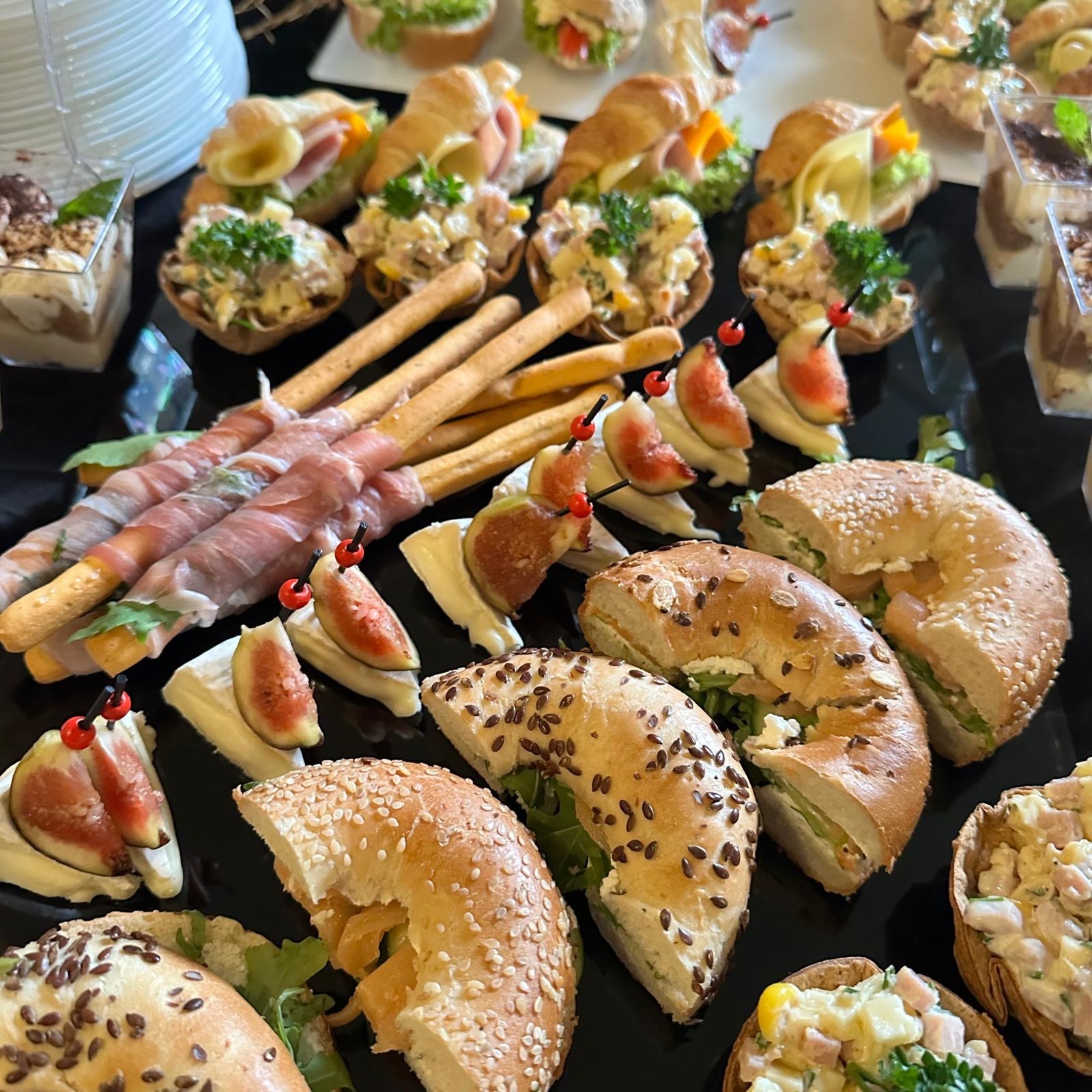 Różnorodny zestaw przekąsek na czarnym talerzu: mini bagietki z łososiem, koreczki z figą i serem, grissini owinięte szynką parmeńską, sałatki w kruchych babeczkach i mini desery w przezroczystych...