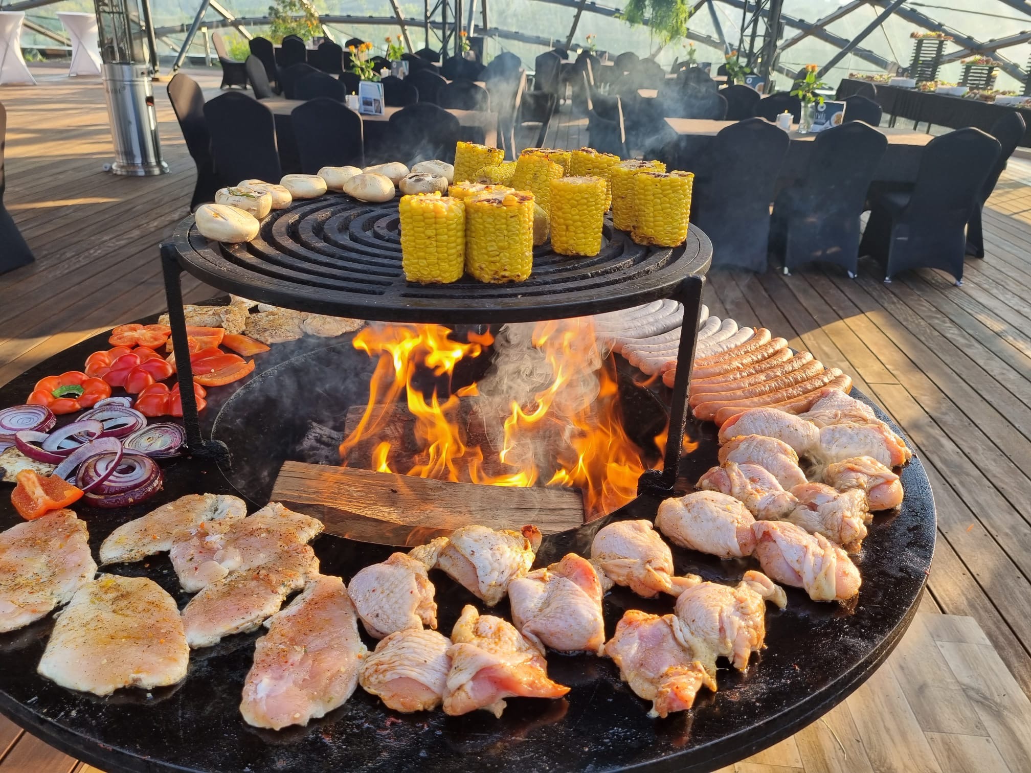 Okrągły grill z płomieniami, na którym znajdują się kukurydza, pieczywo, papryka, cebula, kiełbaski i surowe mięso, w tle stoły z czarnymi krzesłami w przeszklonej przestrzeni.