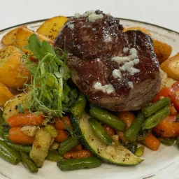 Soczysty stek wołowy z serem, podany na talerzu z pieczonymi ziemniakami i warzywami: fasolką szparagową, marchewką, cukinią i pomidorami.