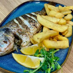 Grilowana ryba dorada z frytkami, plastrami cytryny i świeżymi ziołami na niebieskim talerzu z brązowym brzegiem, ułożona na drewnianym stole.