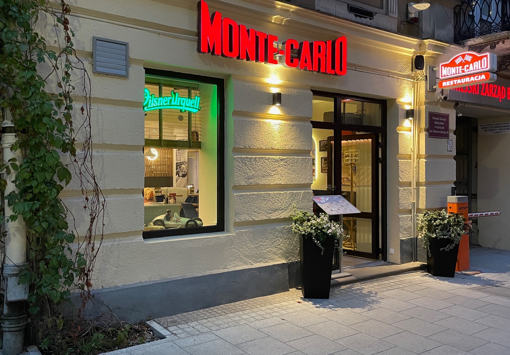 Fasada restauracji Monte-Carlo w Kielcach, z neonowym szyldem i oknem, w którym widoczny jest model samochodu wyścigowego. Menu wystawione przed wejściem, donice z kwiatami po obu stronach.